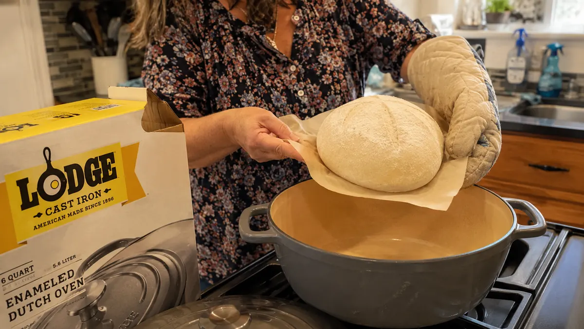 A hand lowering a ball of raw bread dough into a preheated enamel Dutch oven using a sling of parchment paper.