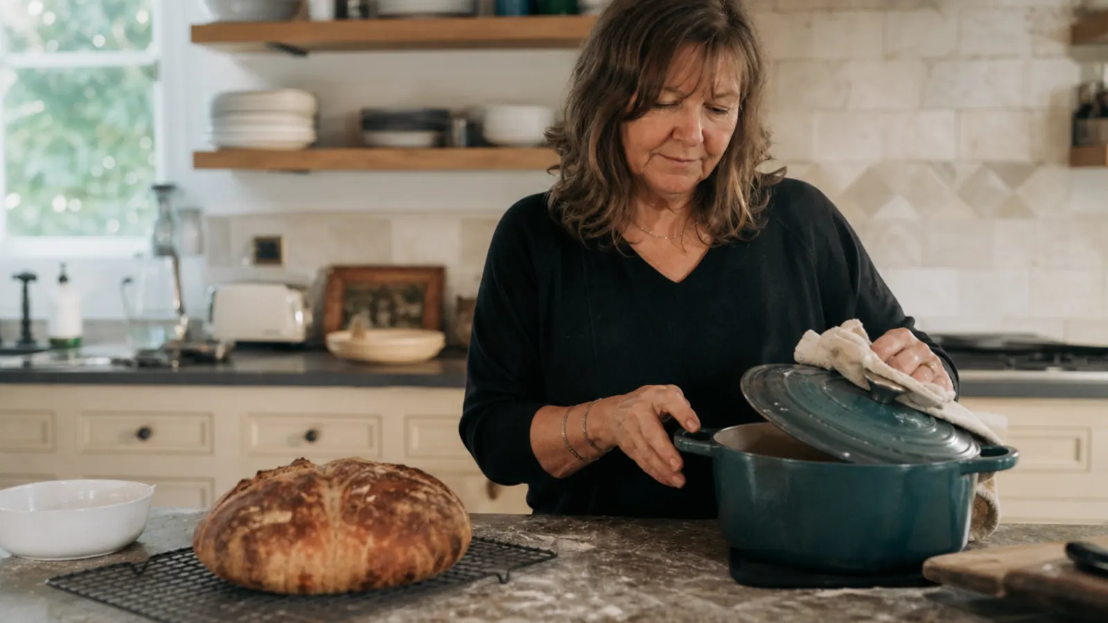 A round crusty loaf of bread cooling on a wire rack next to an open teal enamel Dutch oven on a kitchen counter.